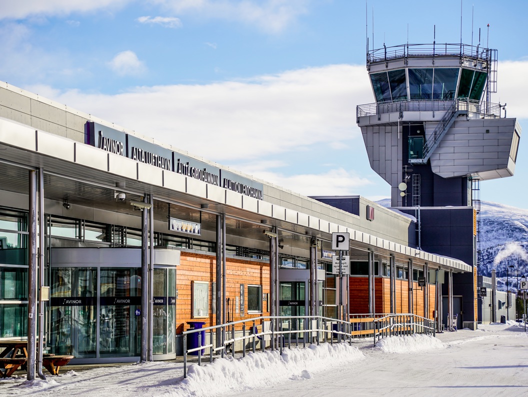 Flyplass med kontrolltårn i vinterlandskap, med skilt for Avinor Alta lufthamn, snødekt bakke og klar blå himmel.