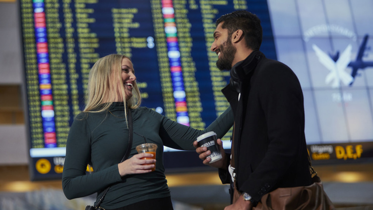 Two people at an airport are smiling and holding drinks, with a departure board visible in the background.