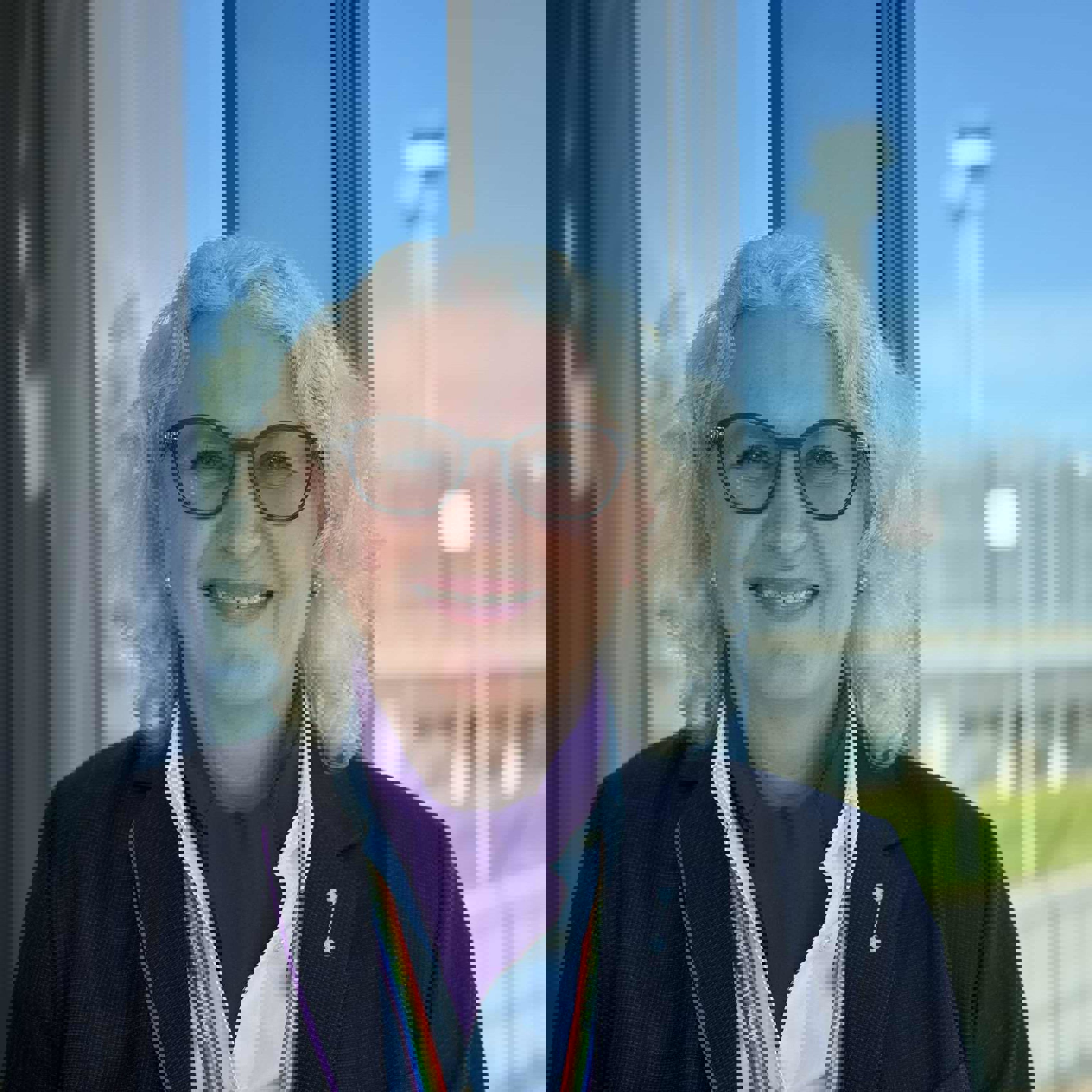 Kvinne med lyst krøllete hår og briller står ved et vindu iført mørk jakke og skjorte, foran en flyplassbakgrunn med klart blå himmel.